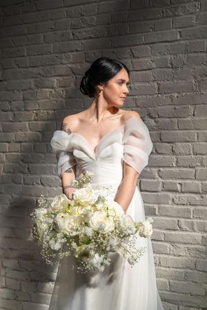 Elegant bride in off-shoulder white gown holding lush white rose bouquet, posing against gray brick wall.