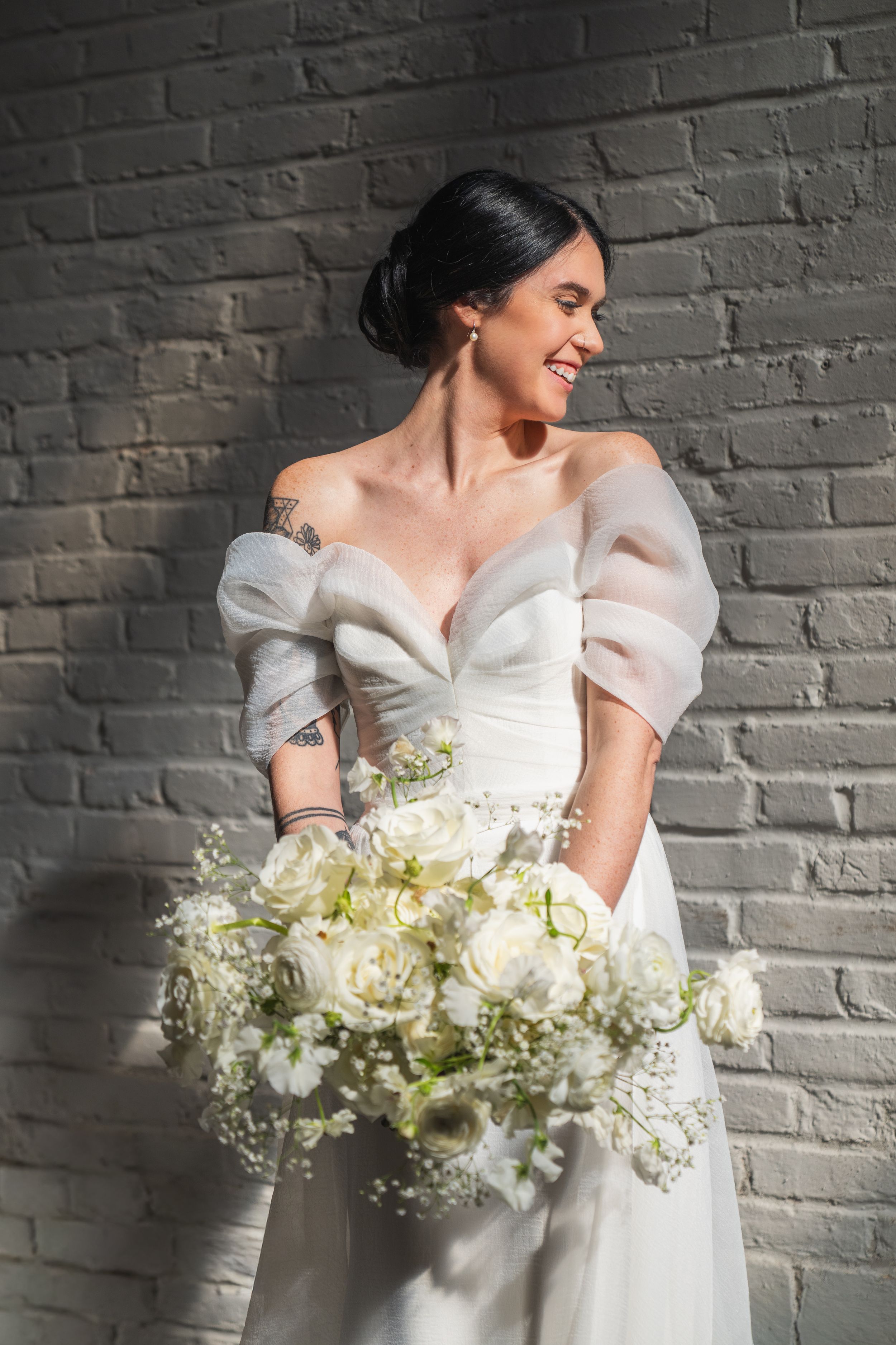 Smiling bride in off-shoulder white gown holding large bouquet of white roses and flowers against brick wall.