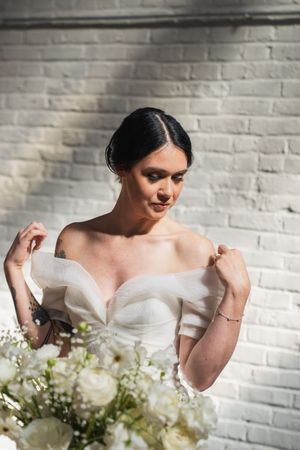 Bride in off-shoulder white gown adjusting dress straps, holding white floral bouquet against brick wall backdrop.
