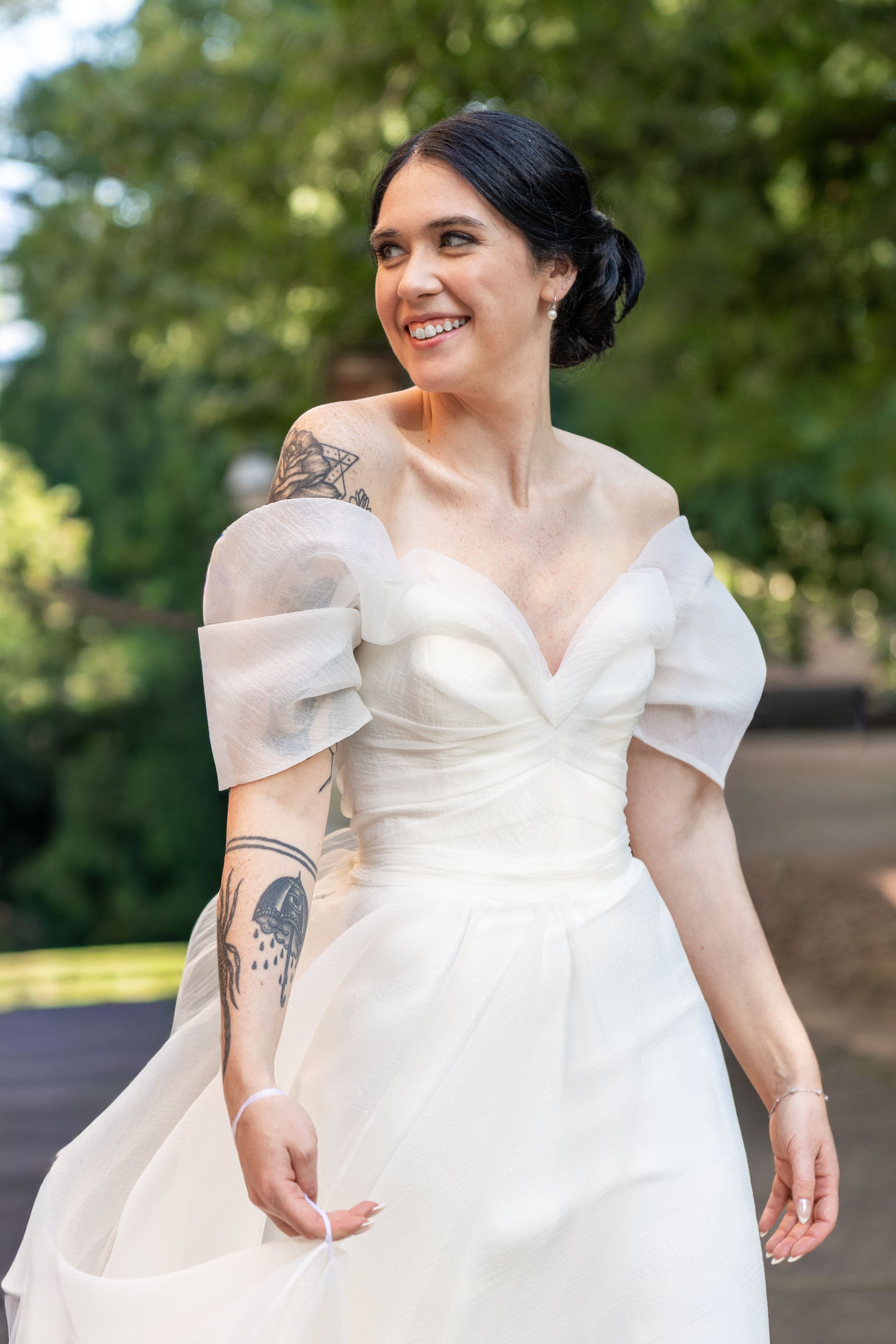 Smiling bride in white off-shoulder gown with sheer sleeves and tattoos, posing outdoors among green trees.
