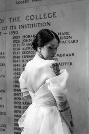 Black and white photo of tattooed woman in white dress posing before college institution wall with engraved names.
