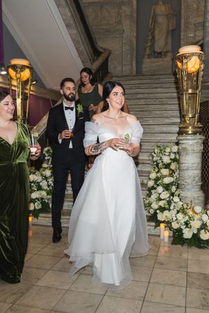Bride in white off-shoulder gown descends grand staircase at elegant wedding venue with floral arrangements.