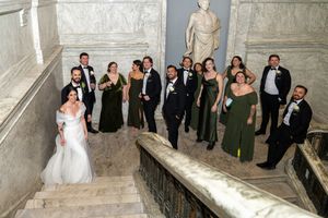 Wedding party poses on grand marble staircase, bride in white gown with groomsmen and bridesmaids in green dresses.