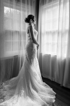 Elegant bride in lace mermaid wedding gown standing by sheer curtained window in black and white photo.