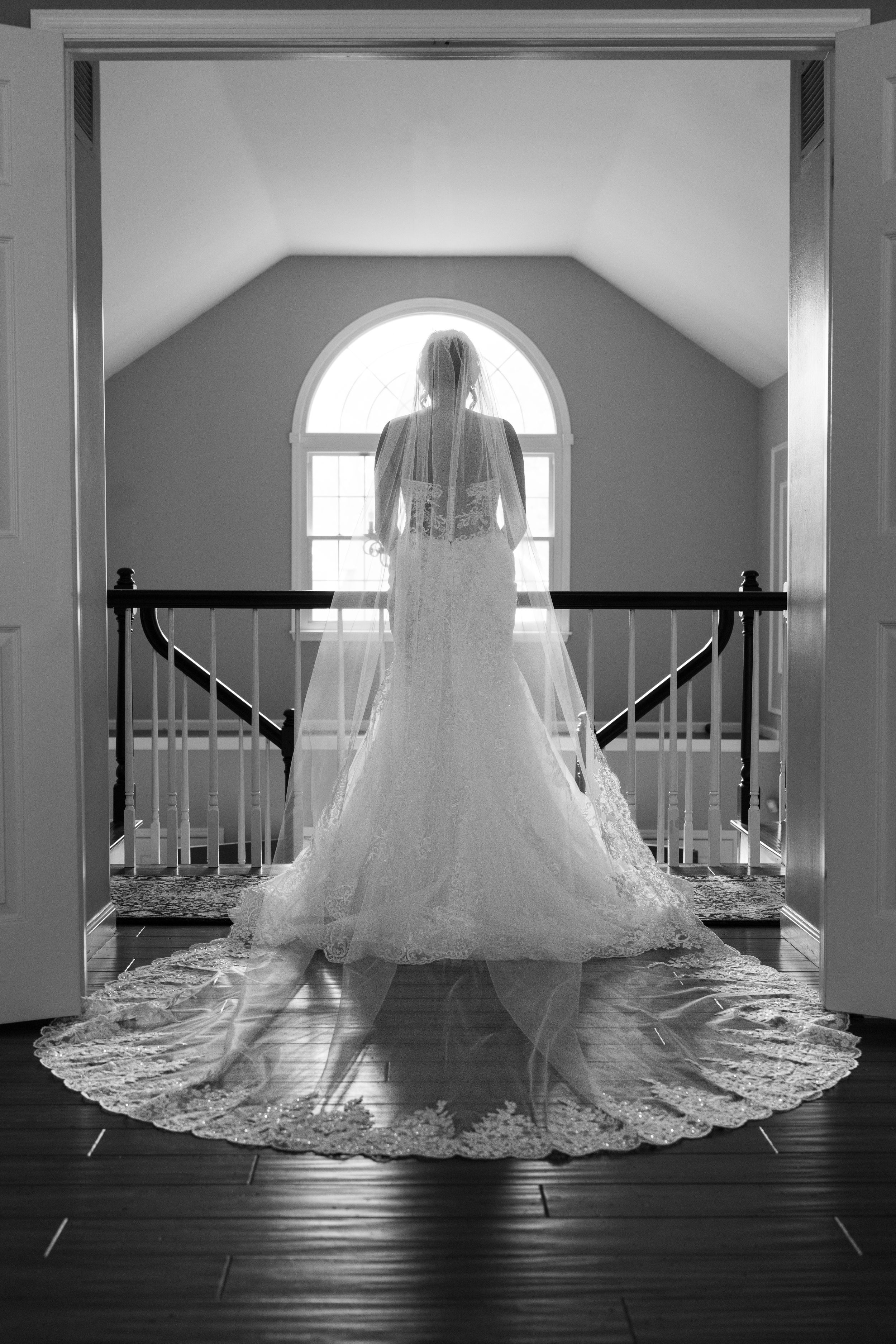 Black and white photo of a bride in a lace mermaid gown with cathedral veil standing before an arched window.