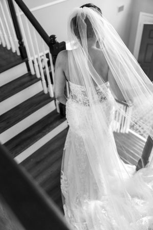 Bride in lace mermaid gown with long veil descending staircase, captured from behind in black and white.