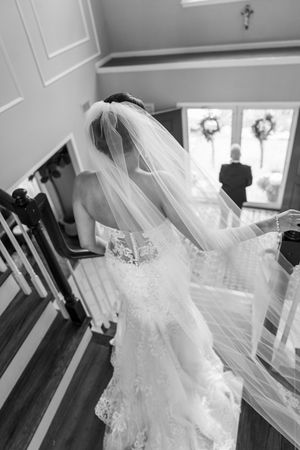 Black and white photo of a bride in lace gown and flowing veil descending a staircase on her wedding day.
