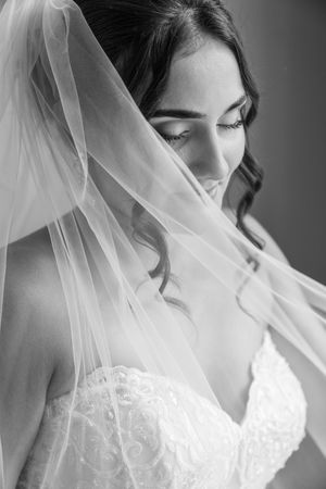 Black and white portrait of a bride with veil draped over her face, wearing a lace wedding dress.