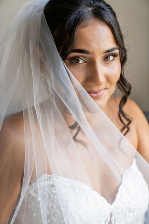 A beautiful bride wearing a white lace gown and sheer veil draped over her shoulders, smiling softly at the camera.
