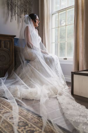 Bride in lace gown and long veil sits by window in soft natural light, smiling on her wedding day.