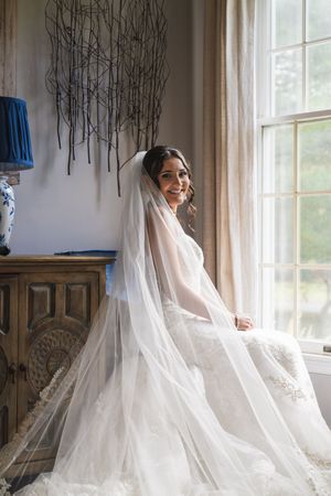 Bride in lace wedding gown and long veil sits by window in elegant room with blue lamp.