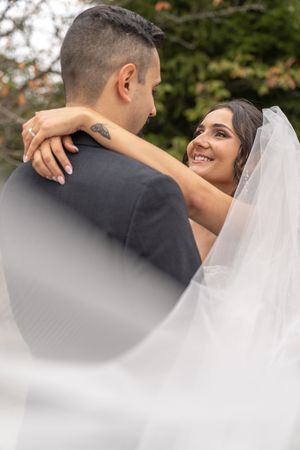 Bride smiling at groom in gray suit, embracing outdoors with flowing veil, lush green trees in background.