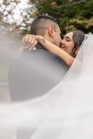 A bride and groom share a romantic kiss outdoors, with her veil flowing in the foreground and lush green trees behind them.