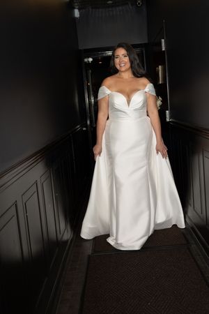 Bride in off-shoulder white satin wedding gown with overskirt walks down dark hallway staircase, smiling elegantly.