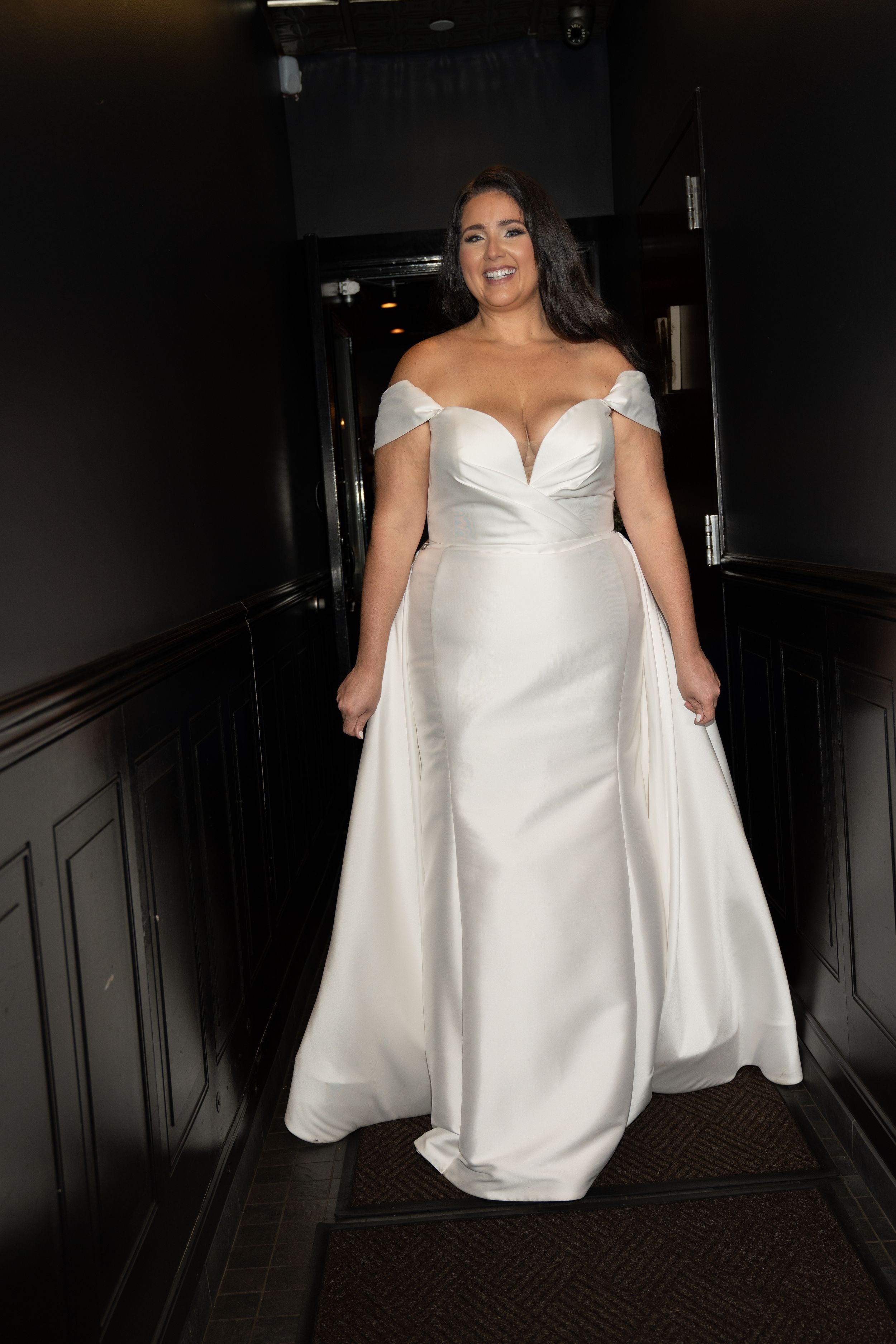 Bride in elegant off-shoulder white satin wedding gown smiling on dark staircase.