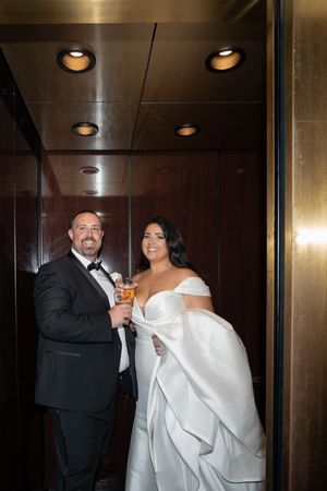 Bride in off-shoulder gown and groom in tuxedo toasting drinks in an elegant elevator.
