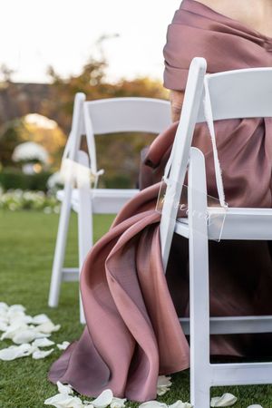 Bridesmaid in dusty rose gown seated on white folding chair at outdoor wedding ceremony with rose petals on grass.