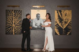 Elegant couple posing in white dress and black tuxedo before gold leaf artwork at a wedding reception.