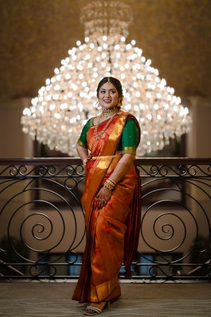 Woman in traditional orange and green saree poses under an elegant crystal chandelier.