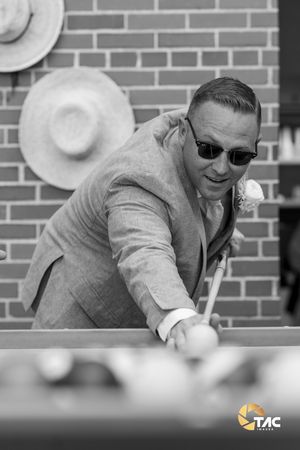 Man in suit and sunglasses playing pool at a wedding reception, leaning over billiard table with cue stick.