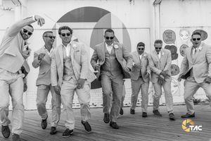 Groomsmen in matching light suits posing playfully on a boardwalk in a stylish black and white wedding photo.