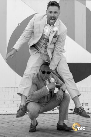 Two men in suits pose playfully outdoors, one crouching while the other leaps over him, in black and white.