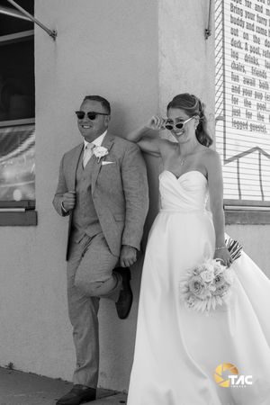 Black and white photo of a bride and groom posing in sunglasses, bride holding a bouquet, leaning against a wall.