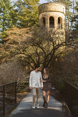 Couple walking on a bridge path beneath a historic stone tower surrounded by autumn trees.