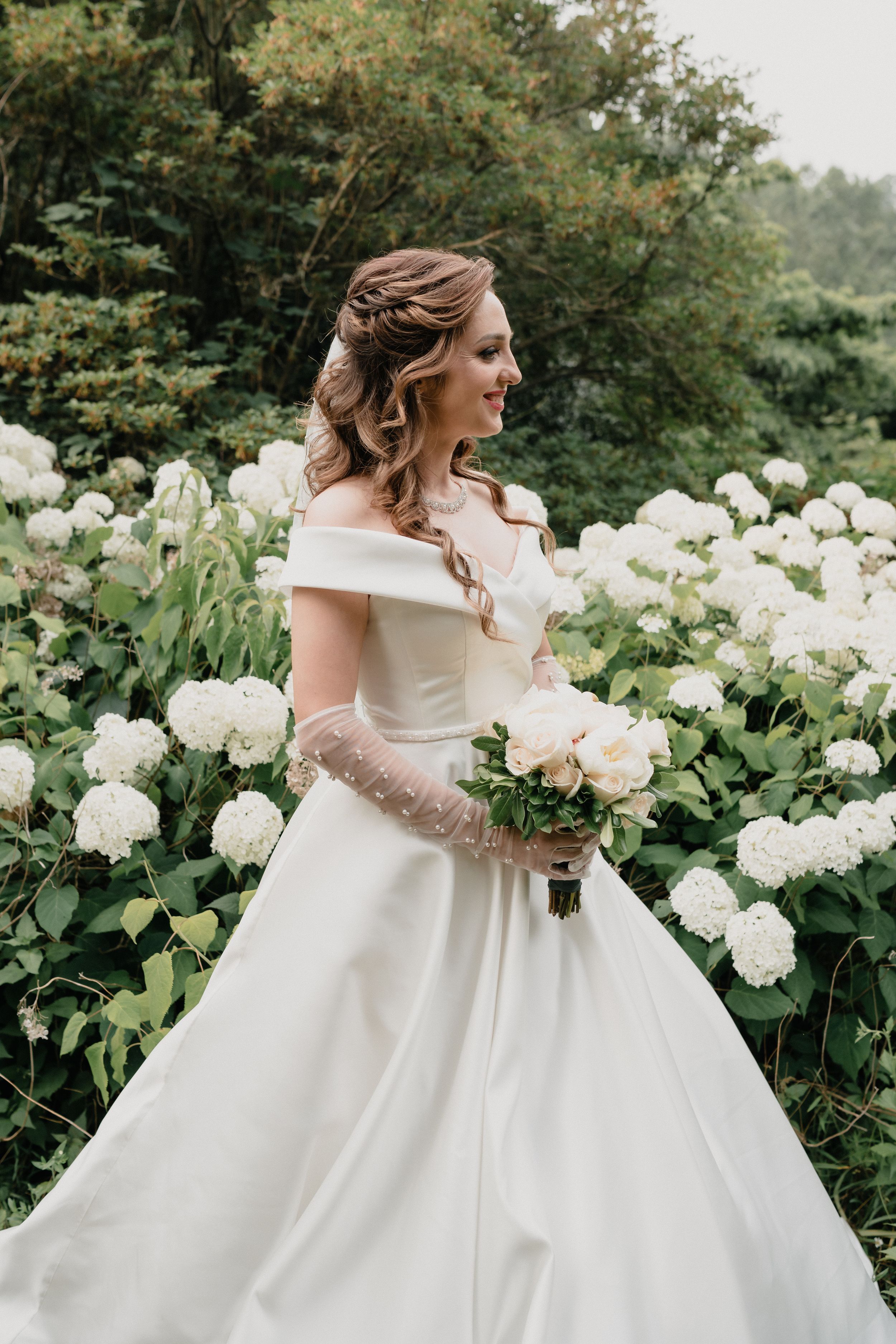 Elegant white wedding dress with full skirt photographed in a garden with white hydrangea flowers.