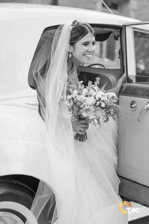 Smiling bride in wedding dress and veil holding bouquet, stepping out of vintage white car in black and white photo.