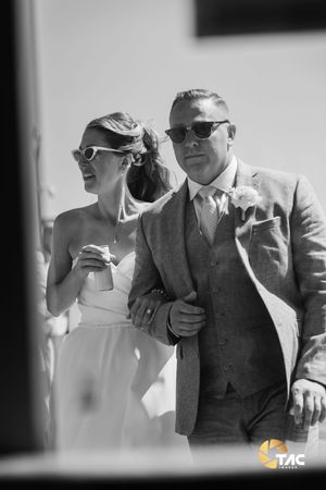 Black and white photo of a bride and groom in sunglasses walking arm in arm at an outdoor wedding ceremony.