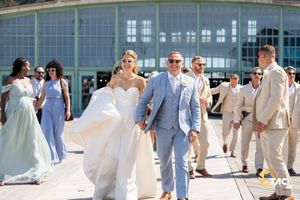 Bride and groom walk hand in hand outdoors with wedding party in blue and beige attire on a sunny day.
