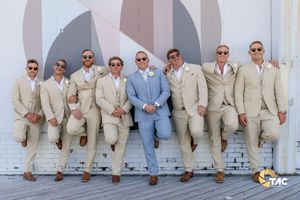 Groom in light blue suit poses with eight groomsmen in tan suits outdoors on a sunny wedding day.