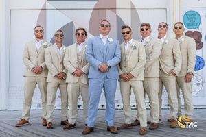 Groom in light blue suit stands with groomsmen in tan suits and sunglasses outdoors at a wedding.