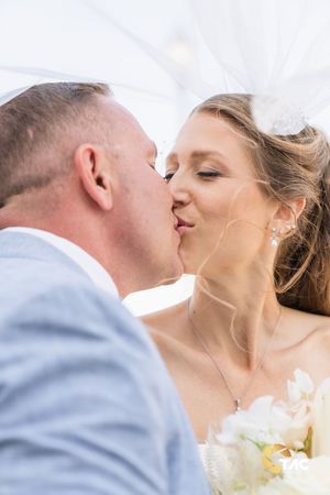 Bride and groom sharing a romantic kiss under a veil on their wedding day, bride holding white flowers.