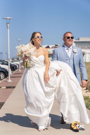 Bride and groom walking outdoors on sunny wedding day, bride holding bouquet and dress, both wearing sunglasses.