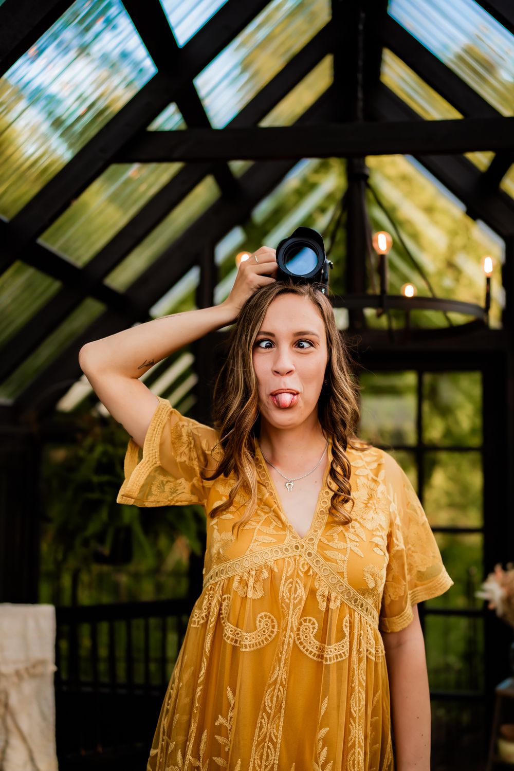 A person in a yellow embroidered dress poses playfully in a rustic greenhouse setting.