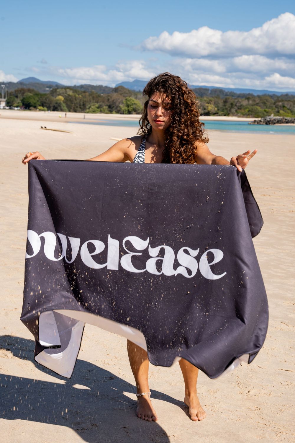 A black beach towel with white text is shown being held up on a sunny beach with mountains in the background.