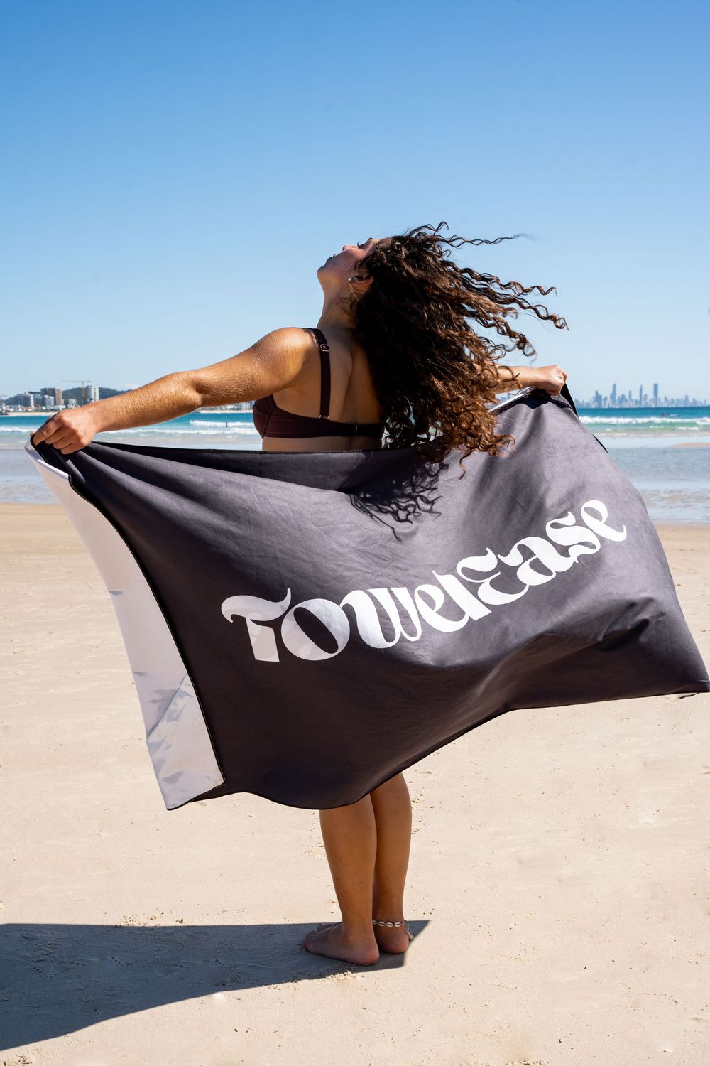 A series of beachside photos showing someone holding and displaying a large black towel with white text against sandy shores.