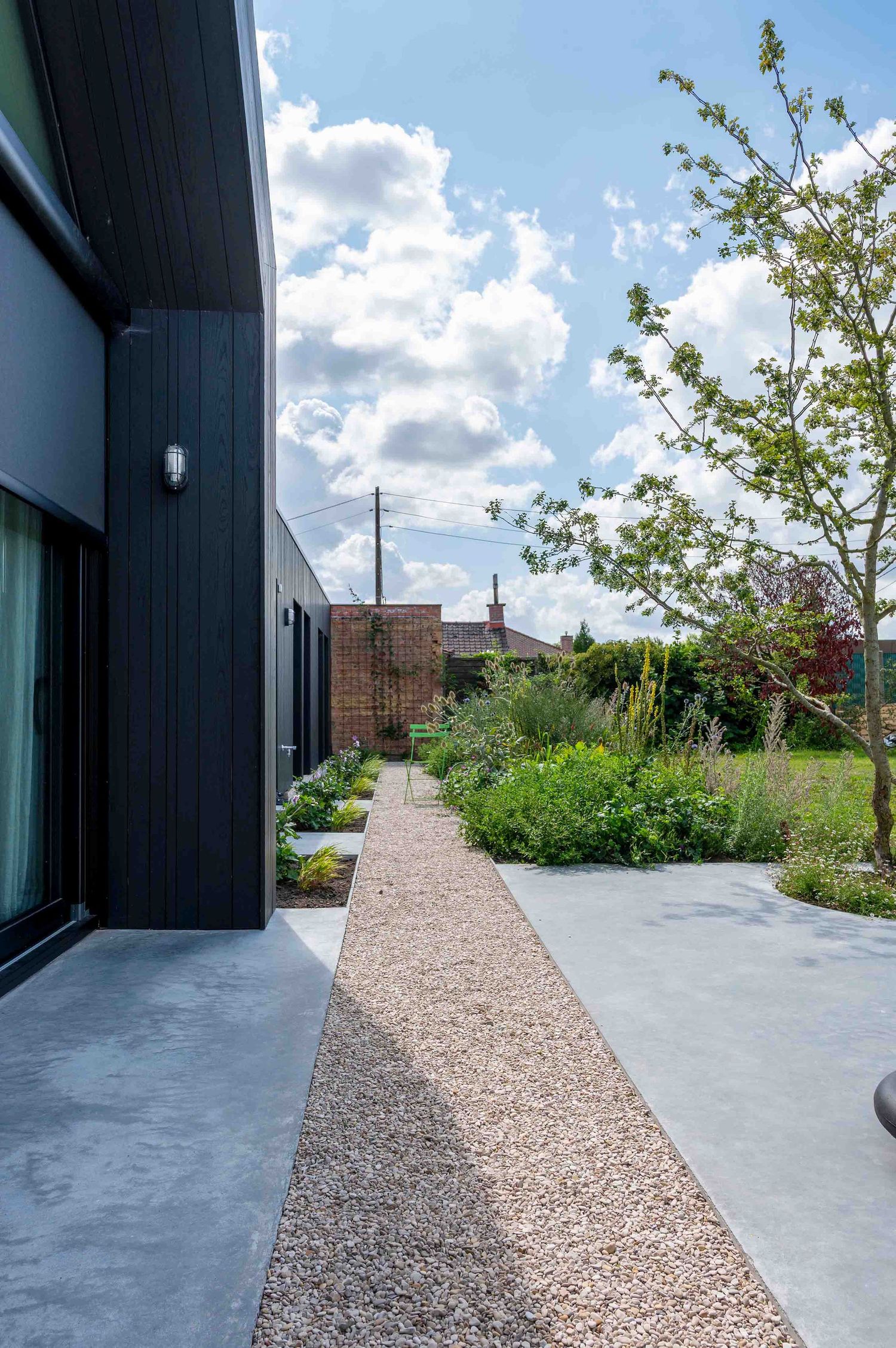 Exterieur van een moderne residentiële woning met een grindpad, een polybeton vloer en beplanting onder een blauwe lucht.