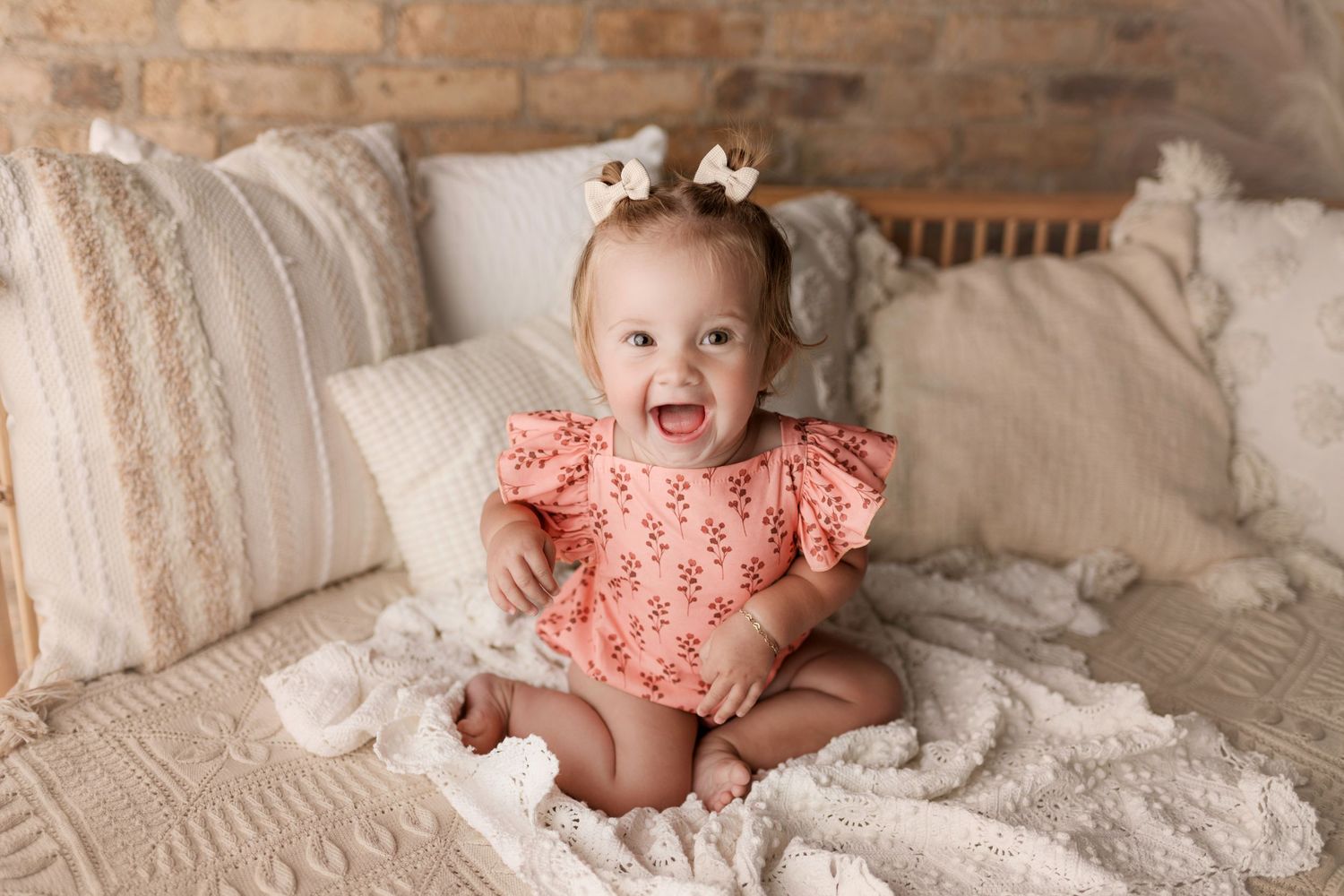 A happy baby girl in a pink floral romper sits laughing on a cozy bed with white textured pillows.