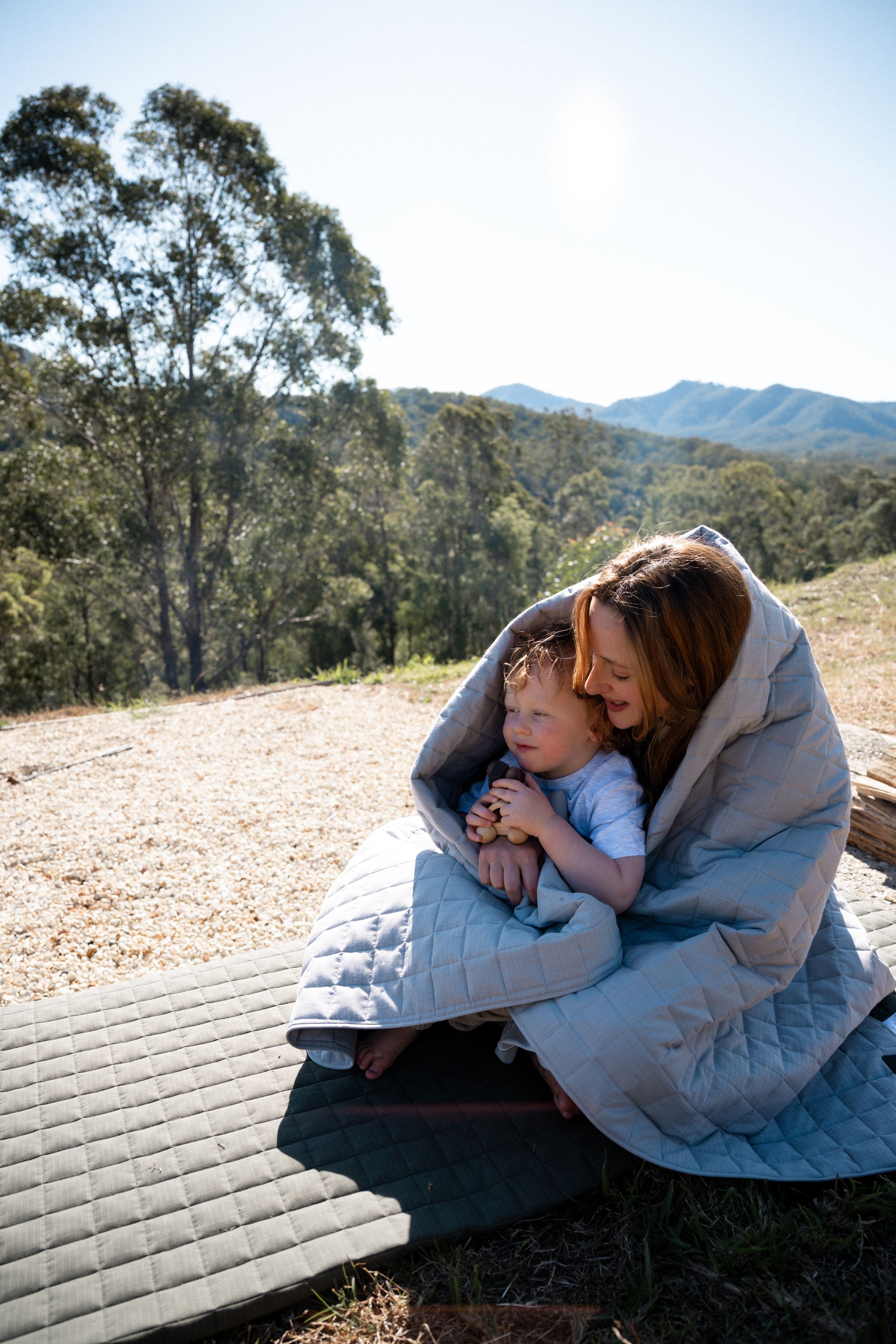Two individuals resting together on a wooden deck overlooking mountains and trees in the background.