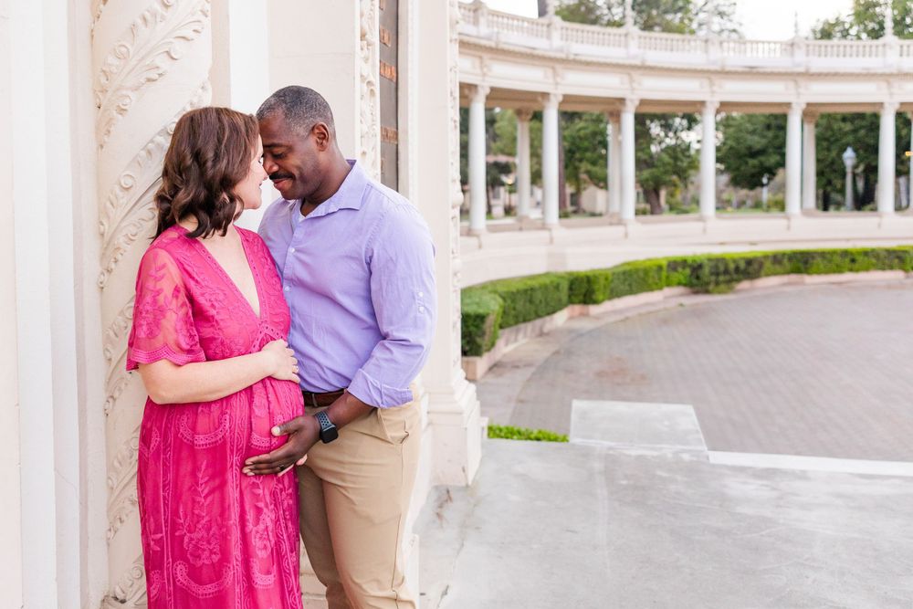 Couple in pink and purple outfits share a tender moment against white pillars in an elegant outdoor setting.