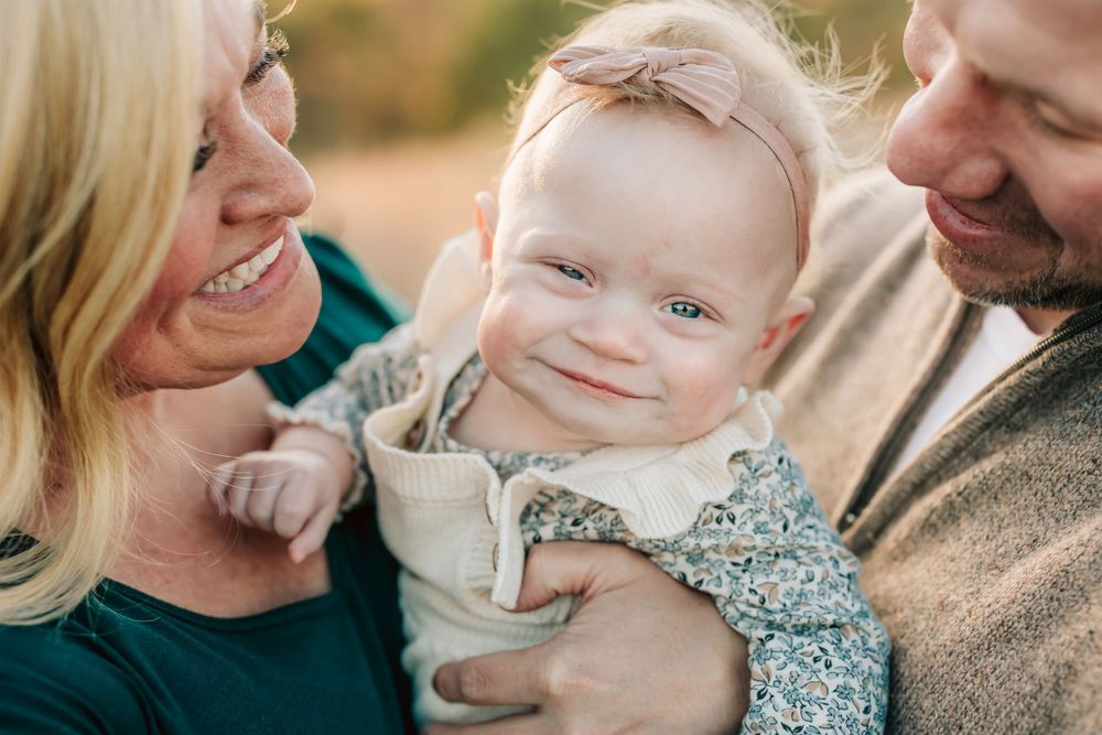 Sweet family portrait with a smiling baby wearing floral print being embraced by loving parents outdoors at sunset.