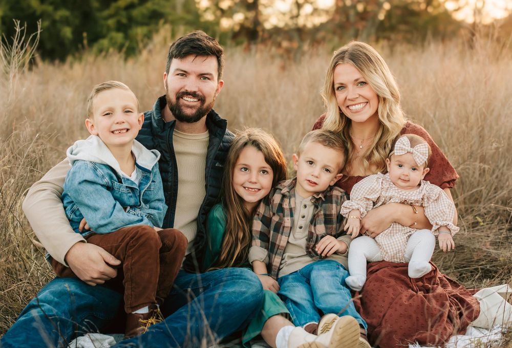 A family of six sits together in a grassy field during golden hour sunset.