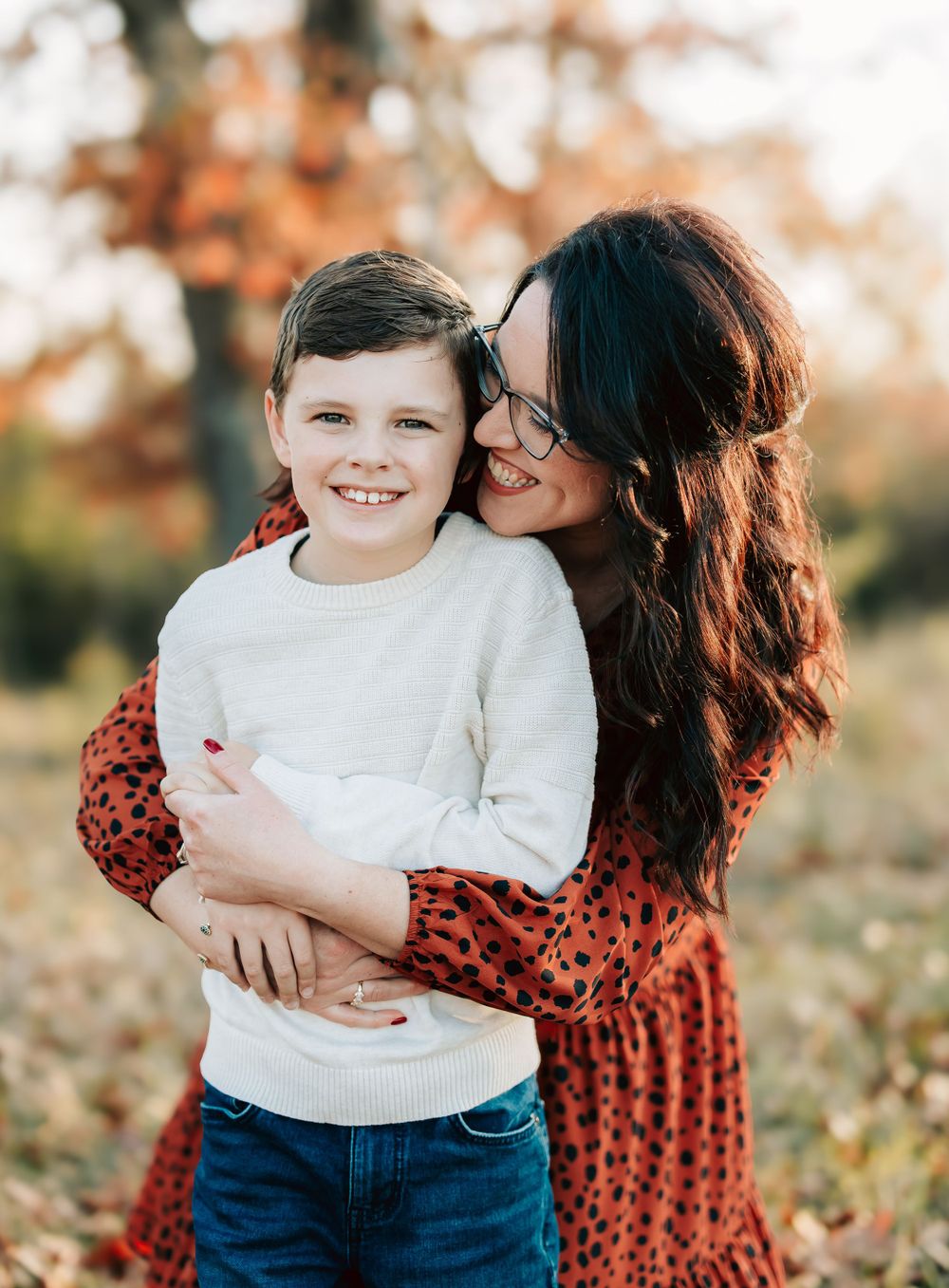 A tender moment captured outdoors with a white sweater and polka dot dress during autumn with fall foliage in background.