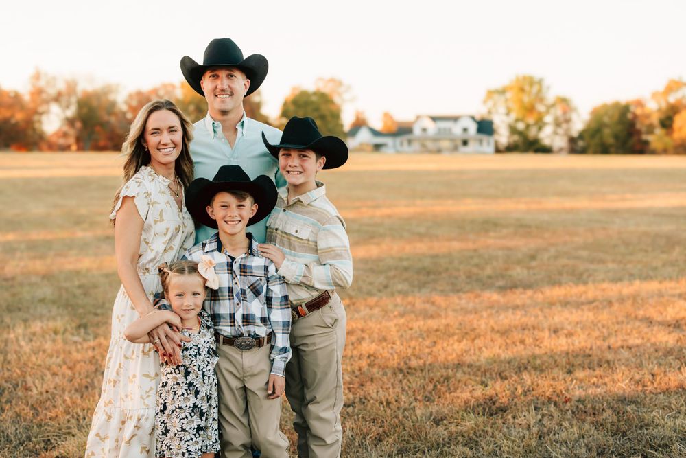 A group wearing cowboy hats stands together in a golden field during sunset wearing western-style attire.