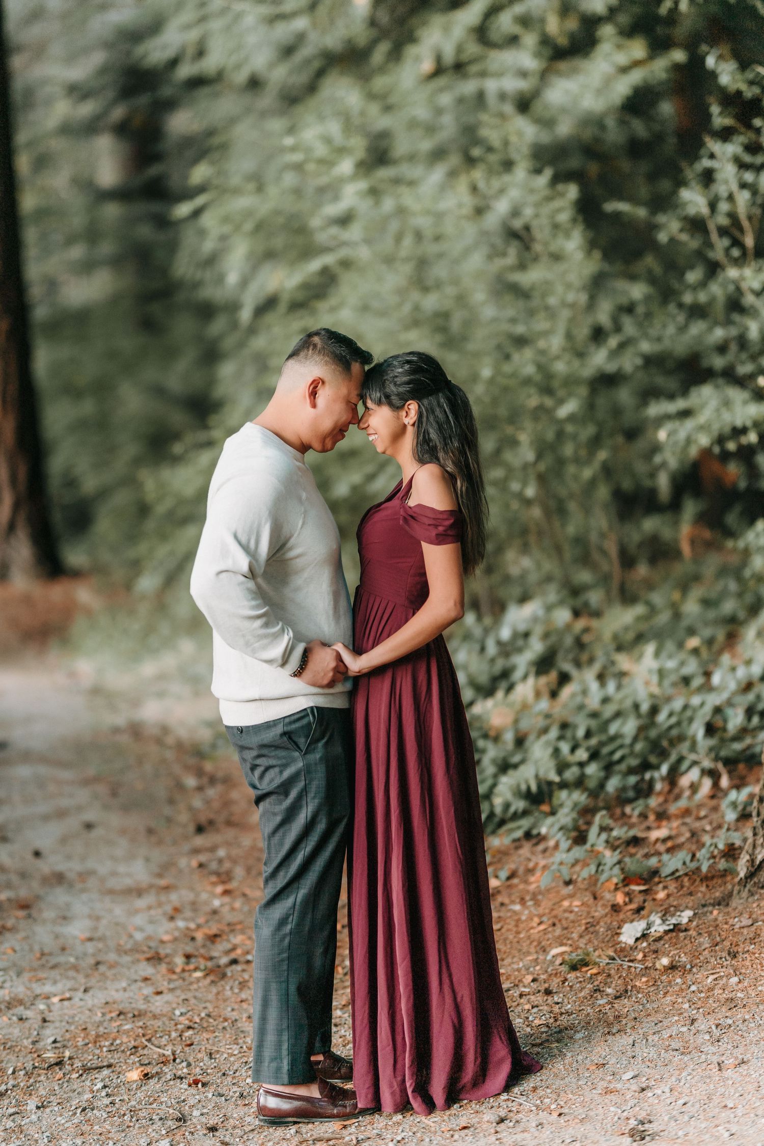 A romantic couple stands intimately on a forest path, with her in a burgundy dress and him in a light sweater.