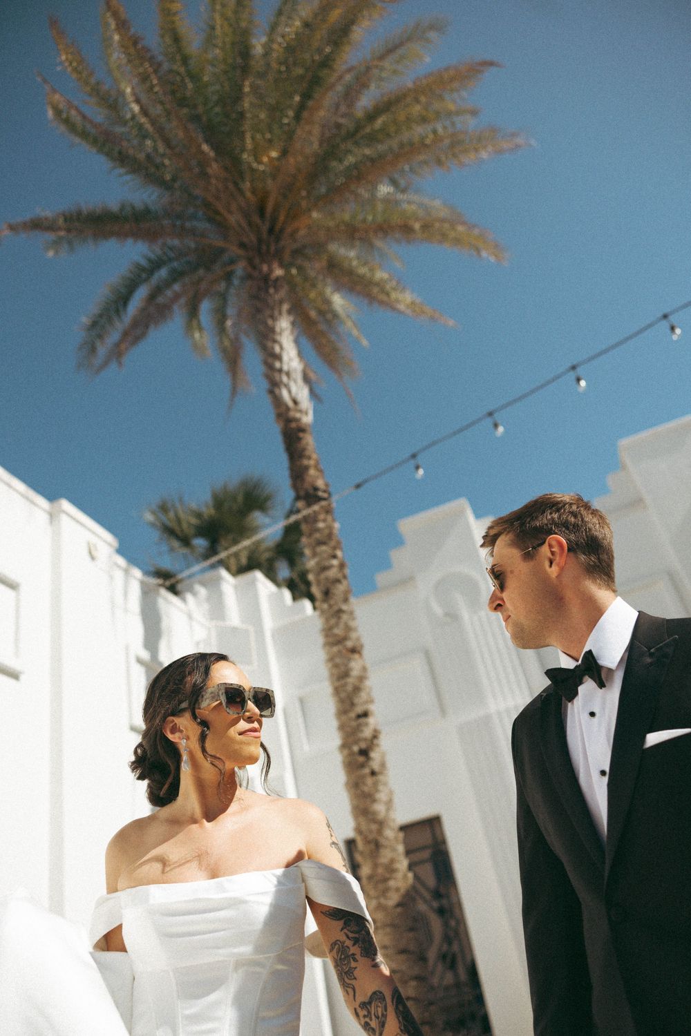 A couple shares a romantic moment under palm trees at an outdoor venue with string lights and white walls.