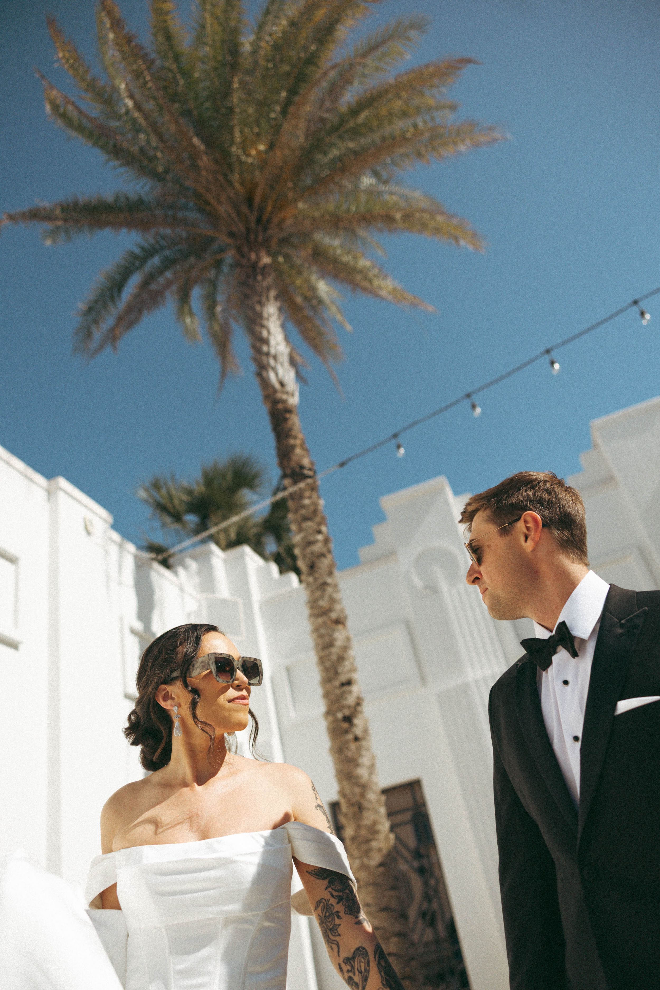 A couple shares a romantic moment under palm trees at an outdoor venue with string lights and white walls.